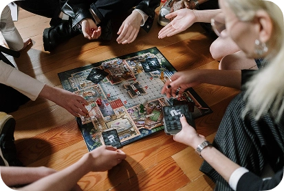 Um grupo de amigos está sentado no chão de madeira a jogar o jogo de tabuleiro Cluedo, com cartas e fichas