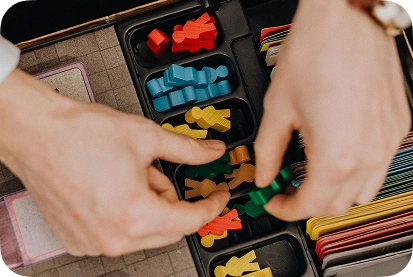 As mãos vão retirando fichas de madeira coloridas com figuras de pessoas do organizador da caixa do jogo de mesa